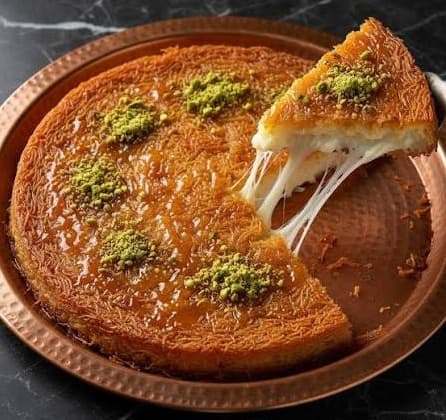 Knafeh ronde sur un plateau en cuivre, une part soulevée laissant filer le fromage, avec de la pistache concassée