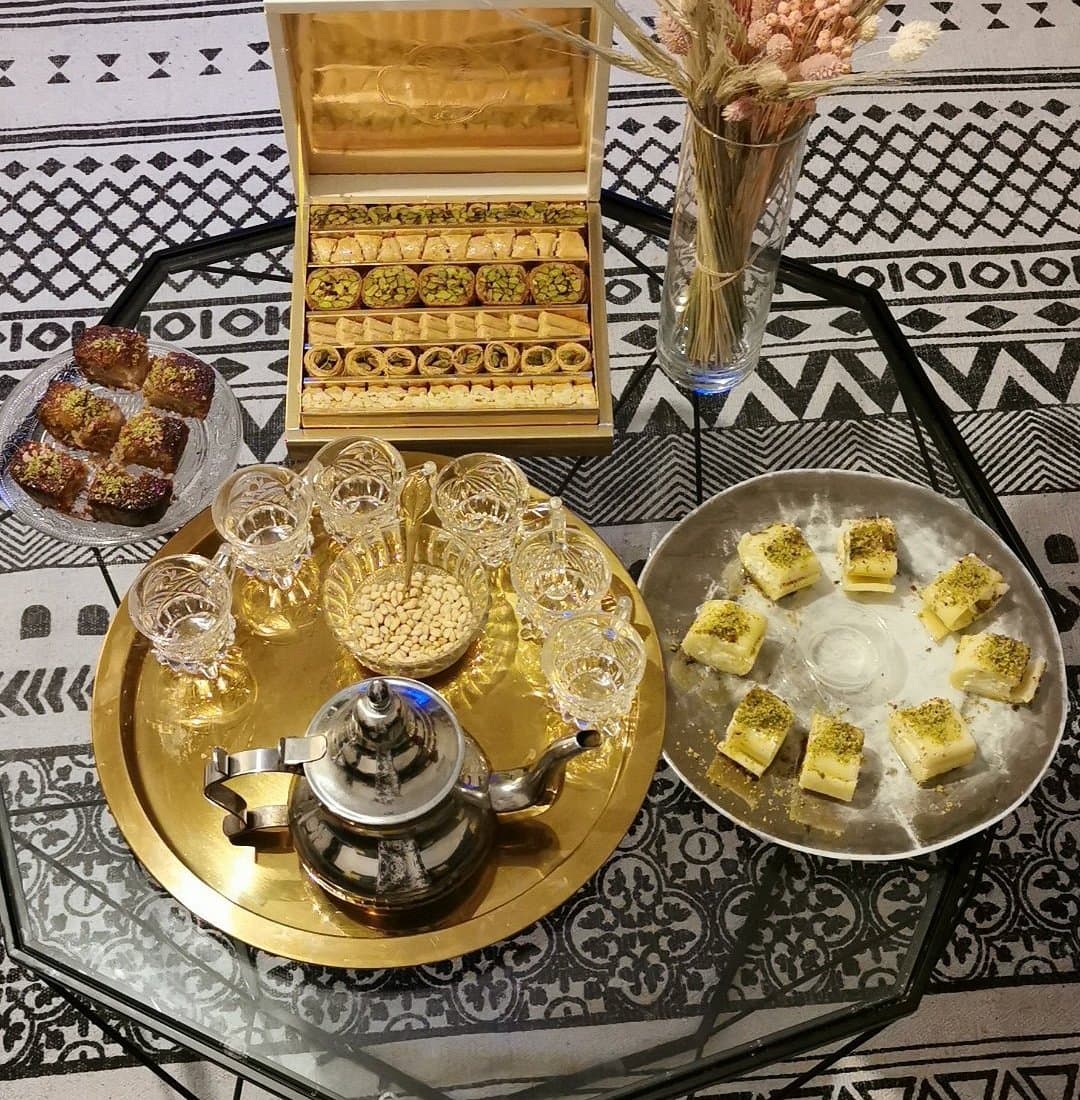 Table dressée vue de dessus : coffret de baklawa ouvert, plateau doré avec théière, verres et pignons de pin, assiette de halawet el jibn à la pistache et pâtisseries